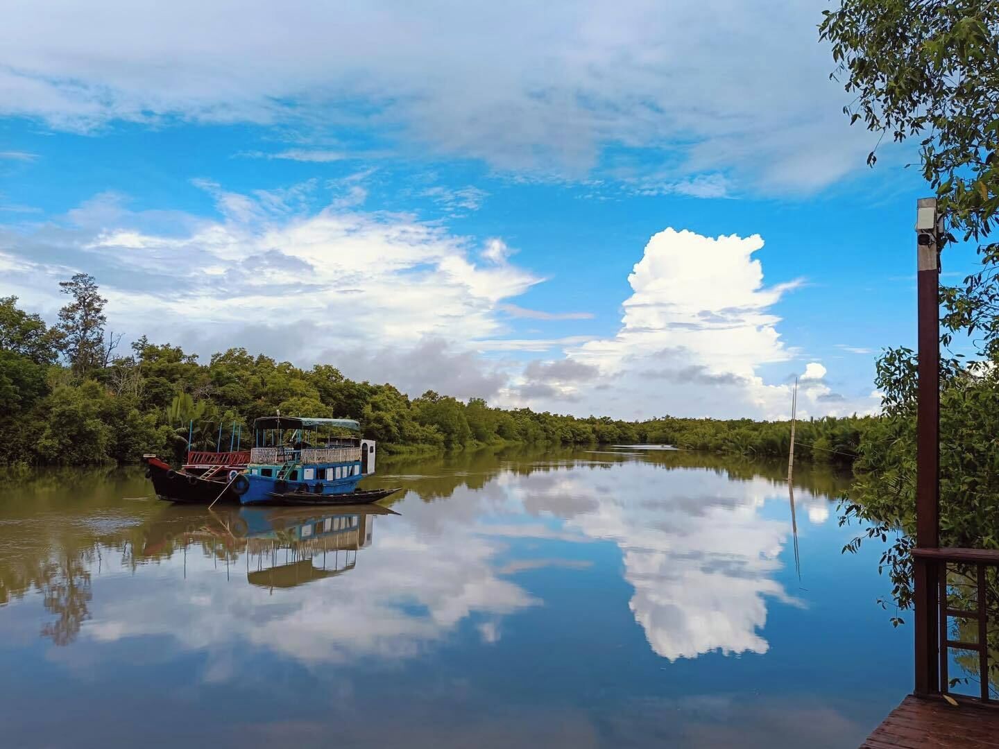 A calm river with a boat docked on the left, surrounded by dense greenery and a clear sky.