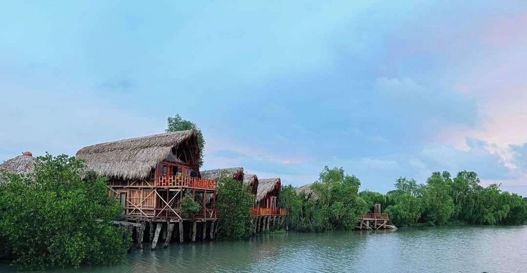 Des bungalows sur pilotis, dotés de toits de chaume et de structures en bois, entourés d’eau et de verdure.