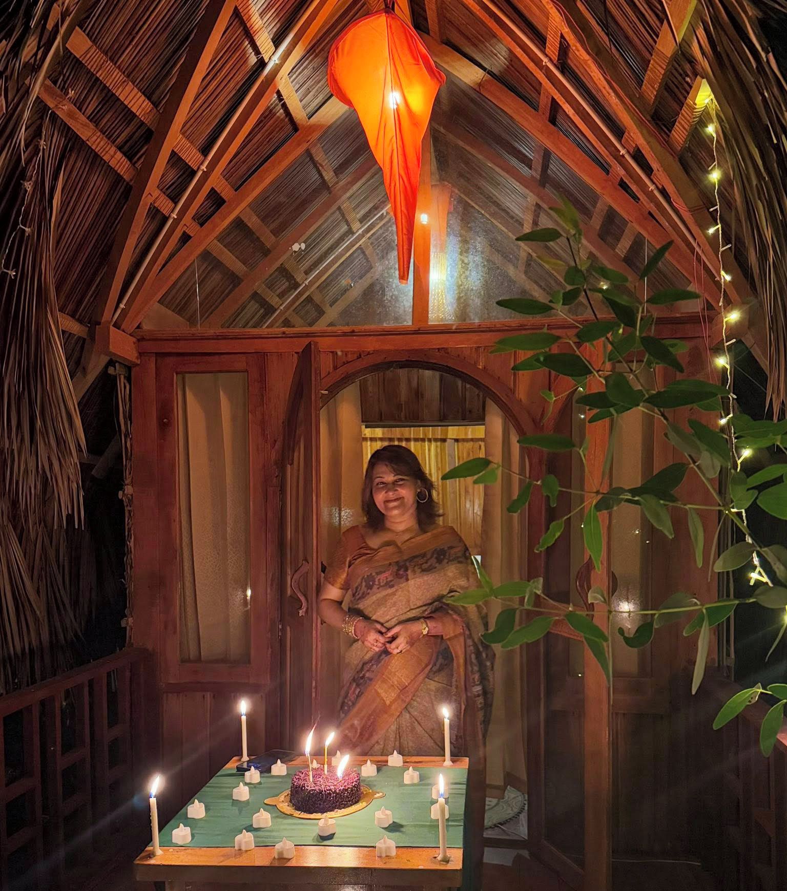 A woman in a traditional sari standing in a wooden hut with a thatched roof, decorated with string lights and candles.