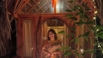 A woman in a traditional sari standing in a wooden hut with a thatched roof, decorated with string lights and candles.