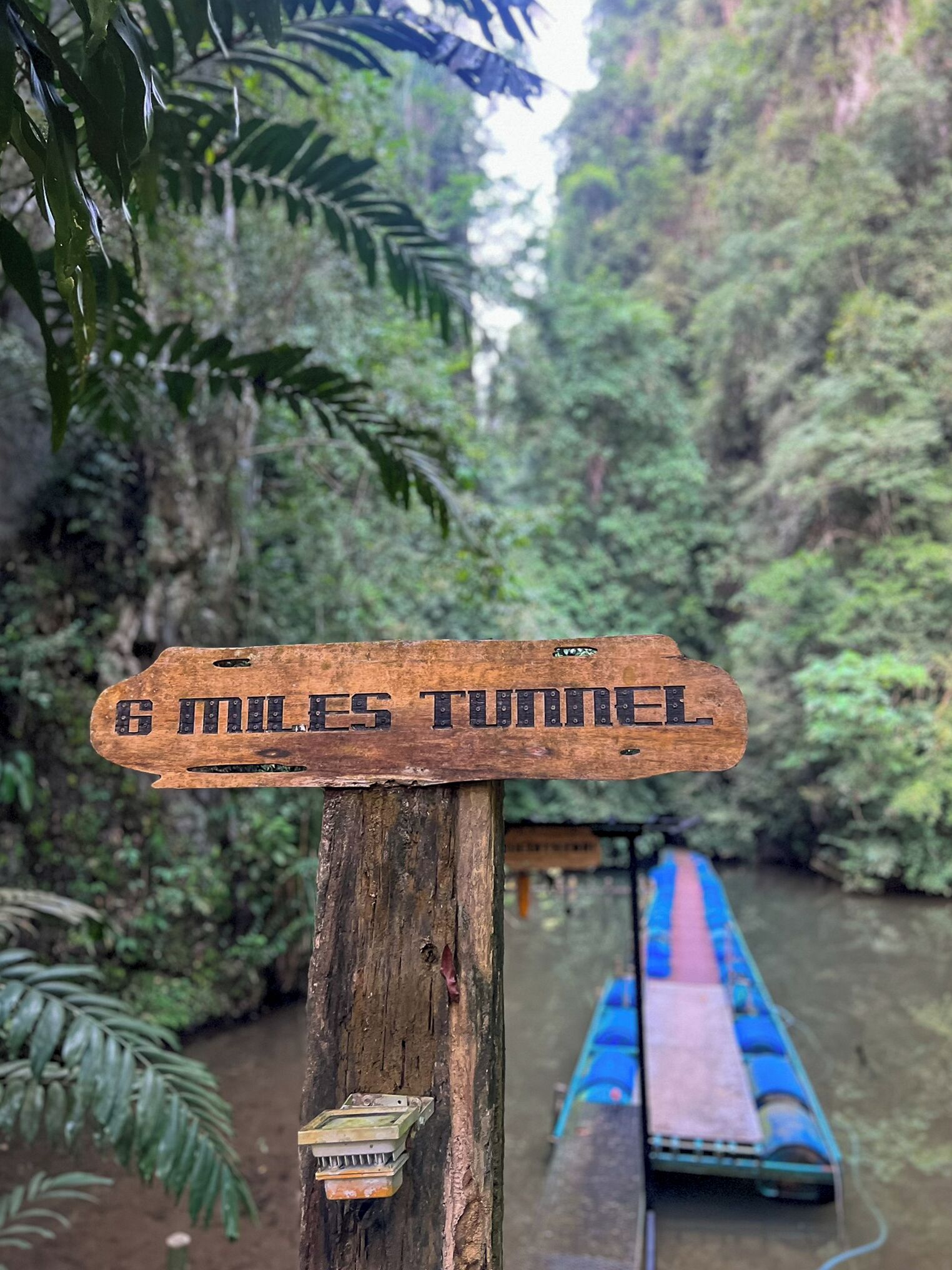 A wooden signpost with the inscription "6 miles tunnel" in a forested area with a river and a boat in the background.