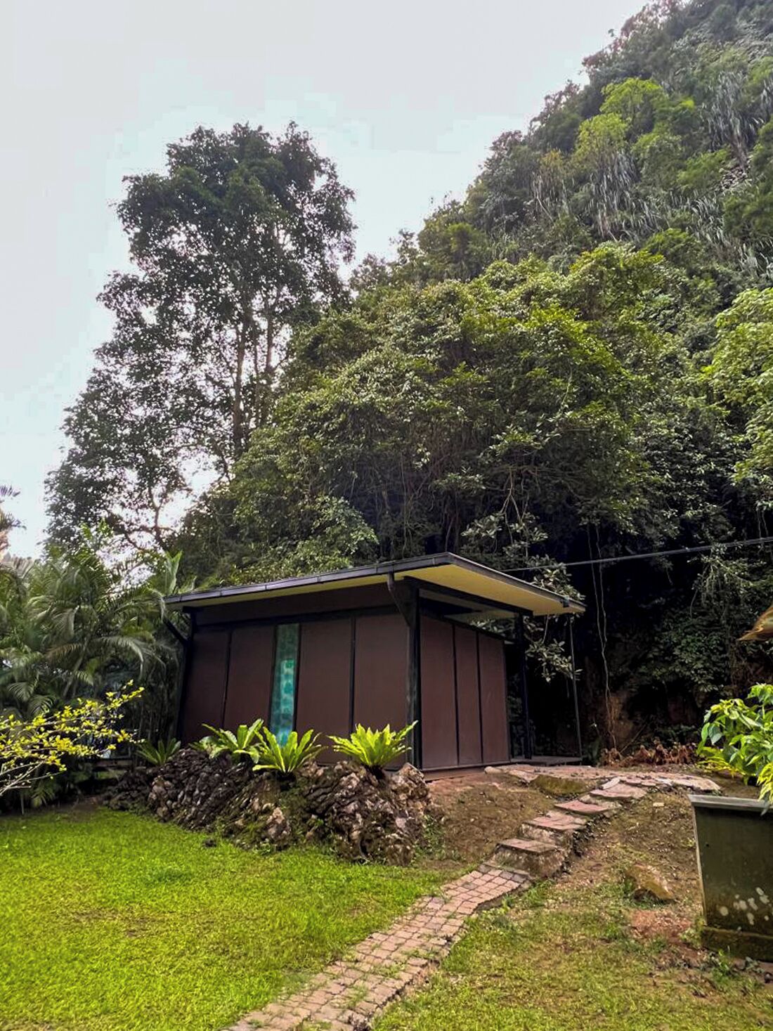 A small, single-story building with a brown exterior and a green door, surrounded by lush greenery and a brick pathway.