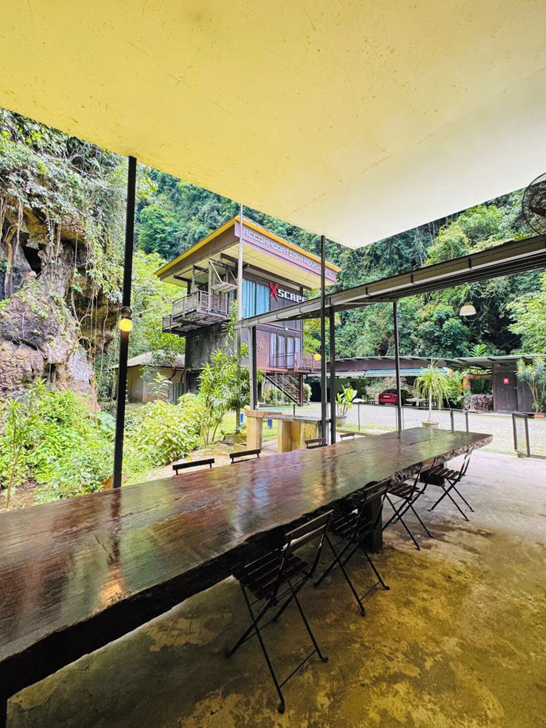A covered outdoor dining area with a long wooden table and chairs, surrounded by lush greenery and a rocky cliff.
