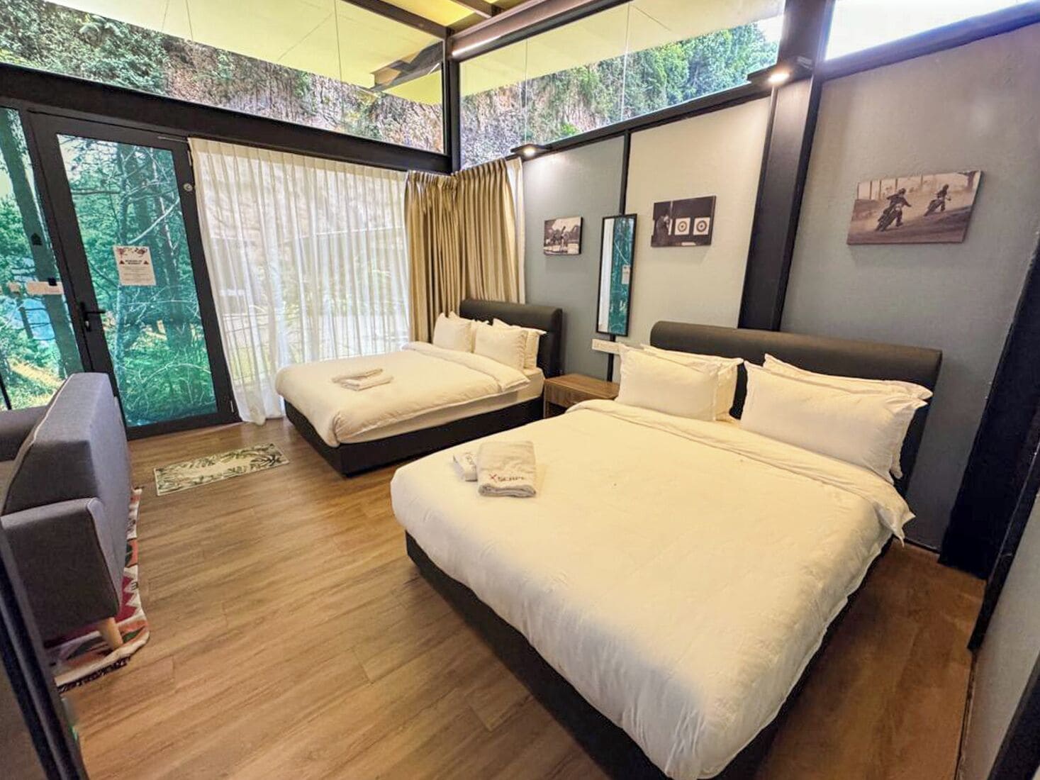 A hotel room with two beds, a large window with curtains, and a view of greenery outside.