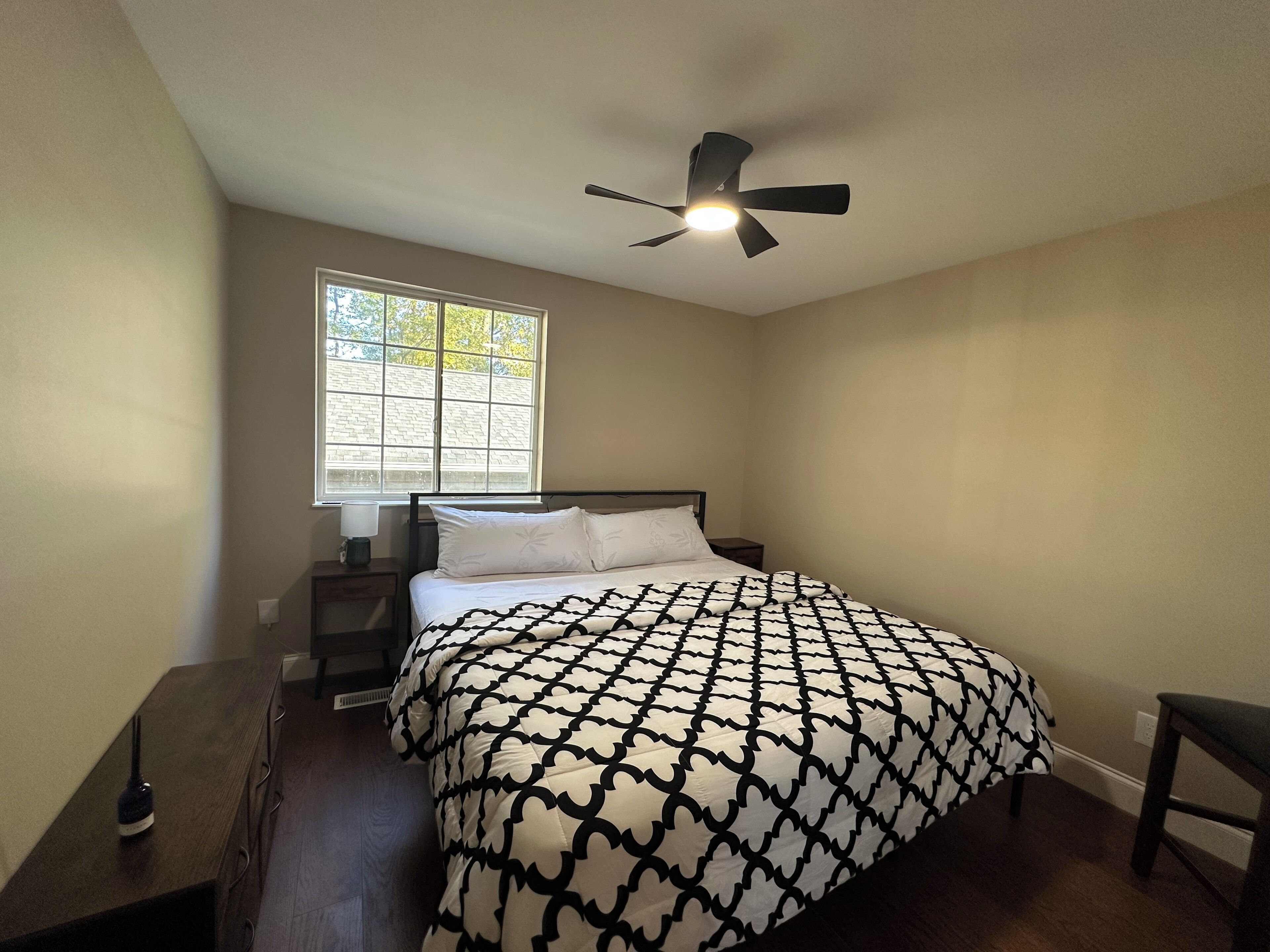 Une chambre à coucher comprenant un lit, une table de chevet, un bureau, une chaise, une fenêtre et un ventilateur de plafond.