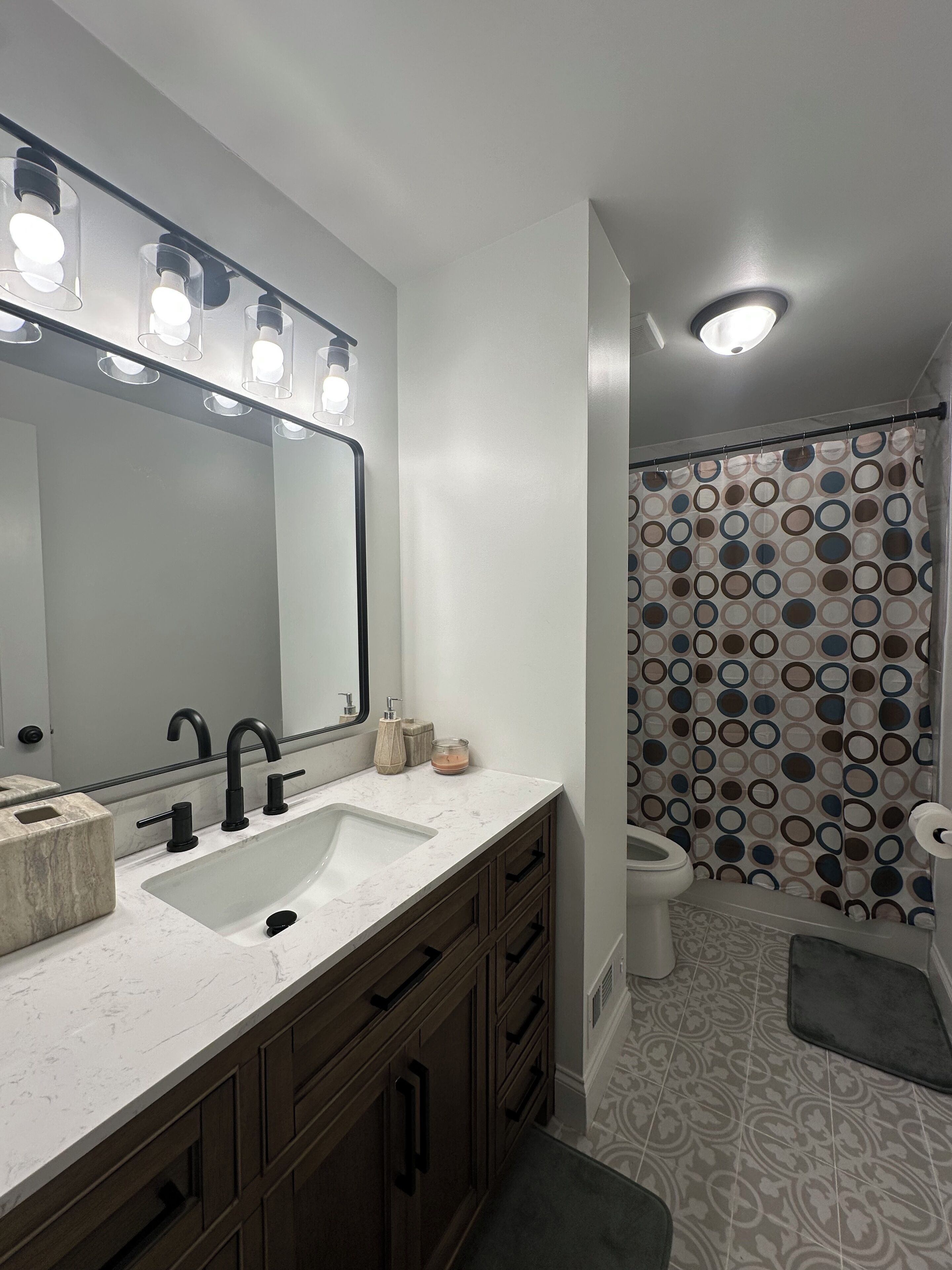 A bathroom with a large mirror, two sinks, a shower curtain with a circular pattern, and a toilet.