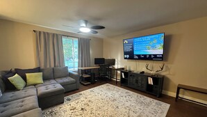 Una sala de estar con un sofá seccional, una alfombra grande, un escritorio con computadora y un televisor de pantalla plana montado en la pared.
