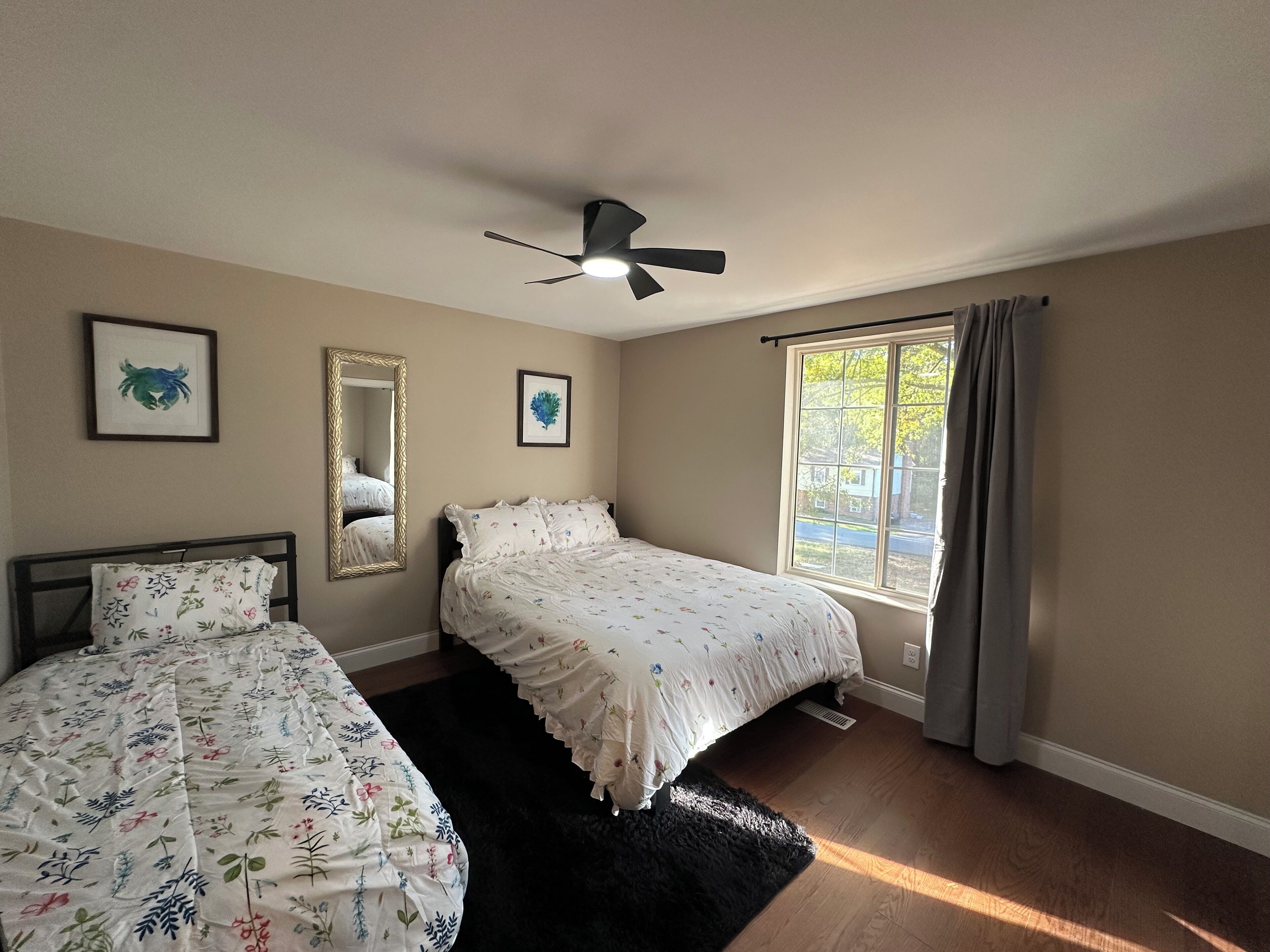 A bedroom with two beds, a ceiling fan, a large window with curtains, and framed artwork on the walls.