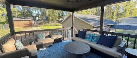 Un área de estar al aire libre techada con muebles de mimbre, una mesa redonda y un tapete azul estampado.