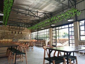 Amplio espacio interior con techos altos, ventanas grandes y plantas colgantes. Mesas y sillas dispuestas para sentarse.