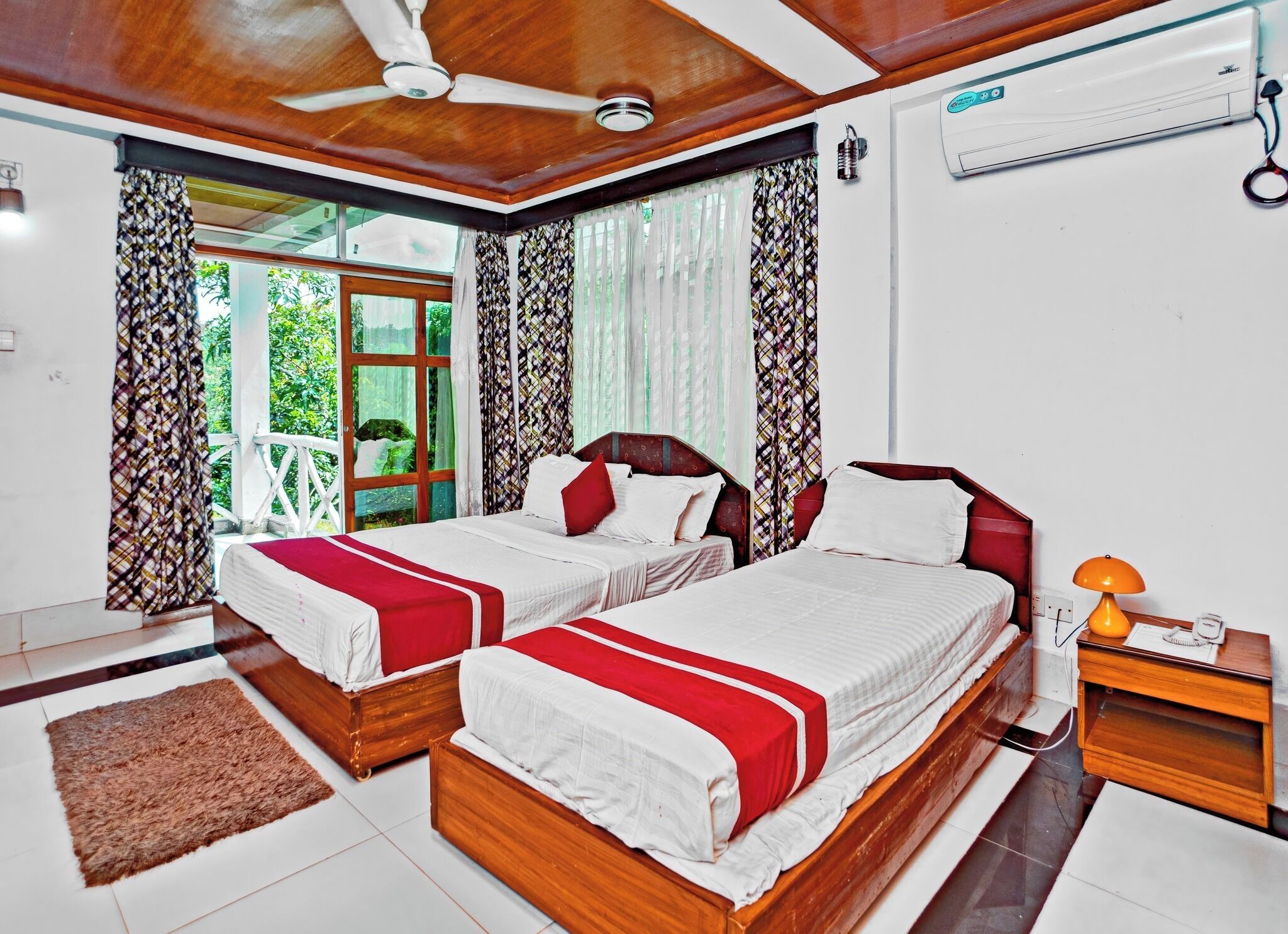A hotel room with two wooden beds, a ceiling fan, an air conditioning unit, and a view of greenery outside.