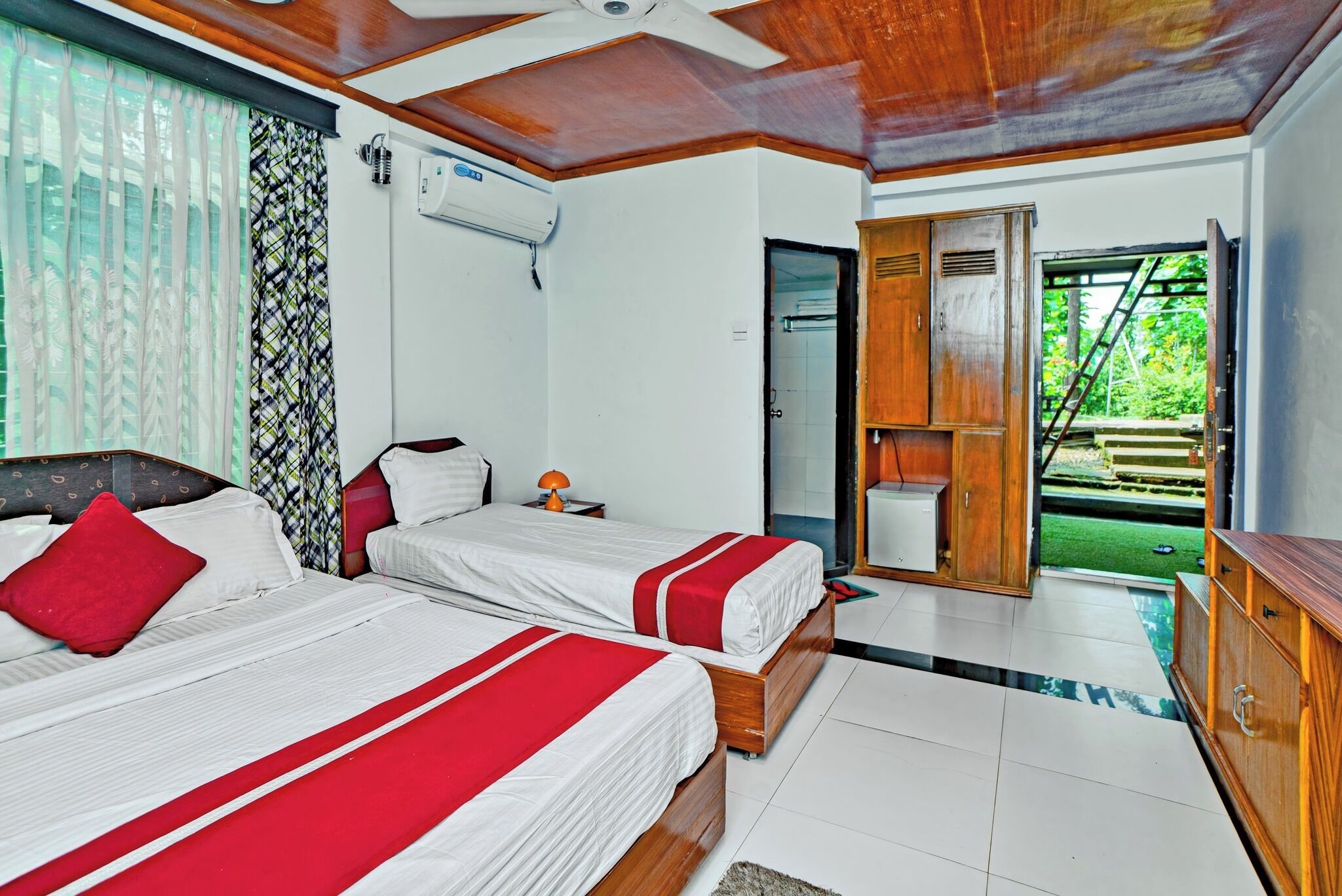 A hotel room with two beds, a wooden wardrobe, a small table with a lamp, and a view of a garden through an open door.