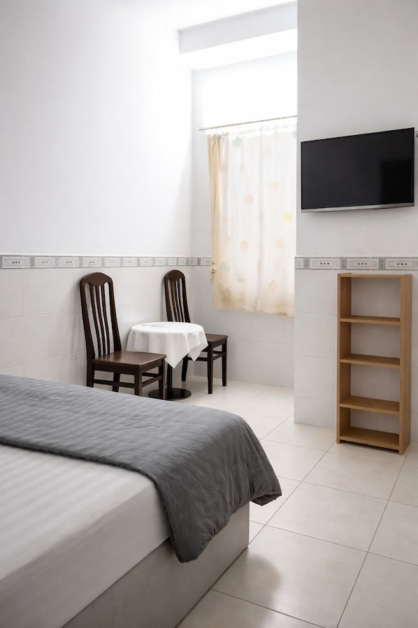 Un dormitorio con cama, una mesita con sillas, un televisor montado en la pared y una ventana con cortina.