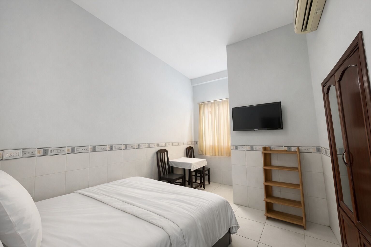 Quarto de hotel com uma cama, uma pequena mesa com cadeiras, uma televisão fixada na parede e uma prateleira de madeira.
