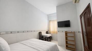 Quarto de hotel com uma cama, uma pequena mesa com cadeiras, uma televisão fixada na parede e uma prateleira de madeira.