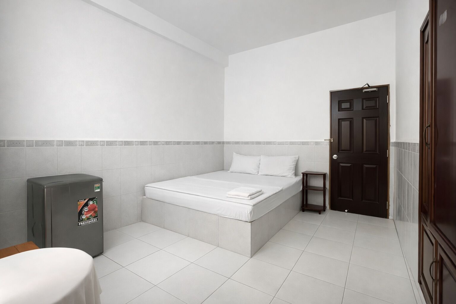 A small, clean room with a bed, a small table, a refrigerator, and a door.