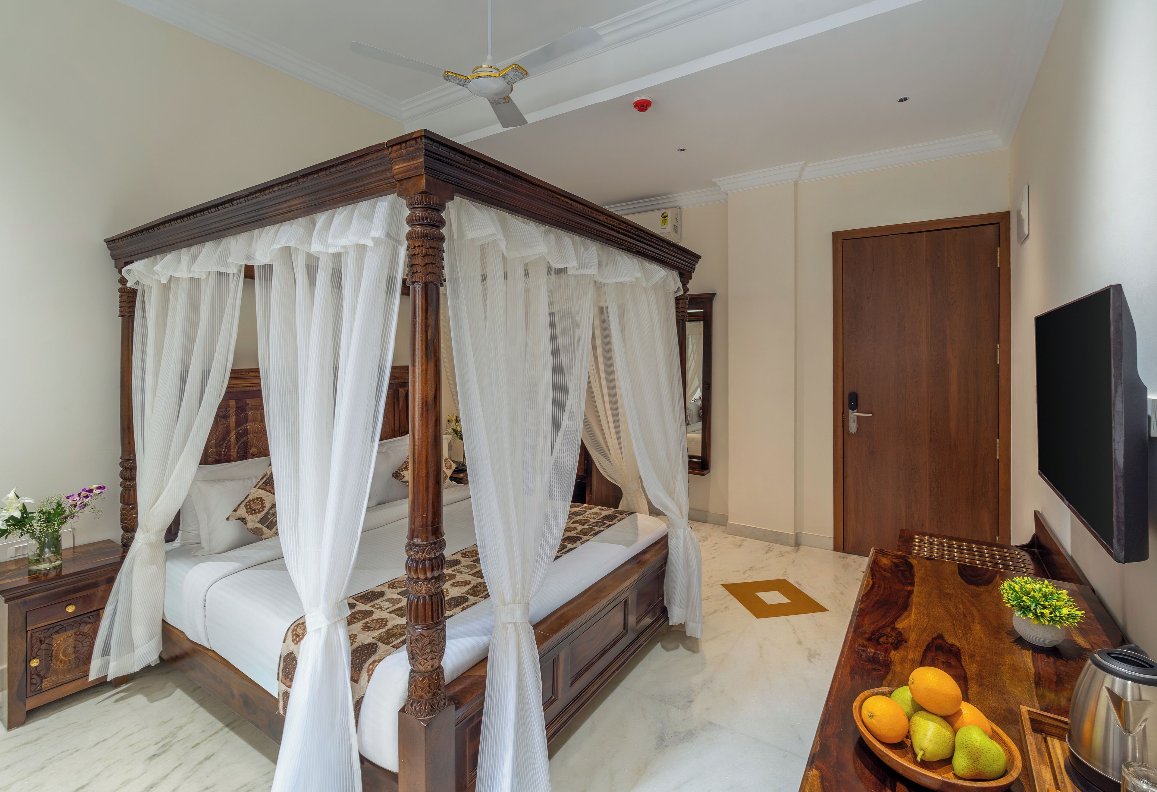 Una cama con dosel y cortinaje, una mesita de noche de madera con un jarrón de flores, un frutero sobre una mesa de madera, y un televisor montado en la pared.