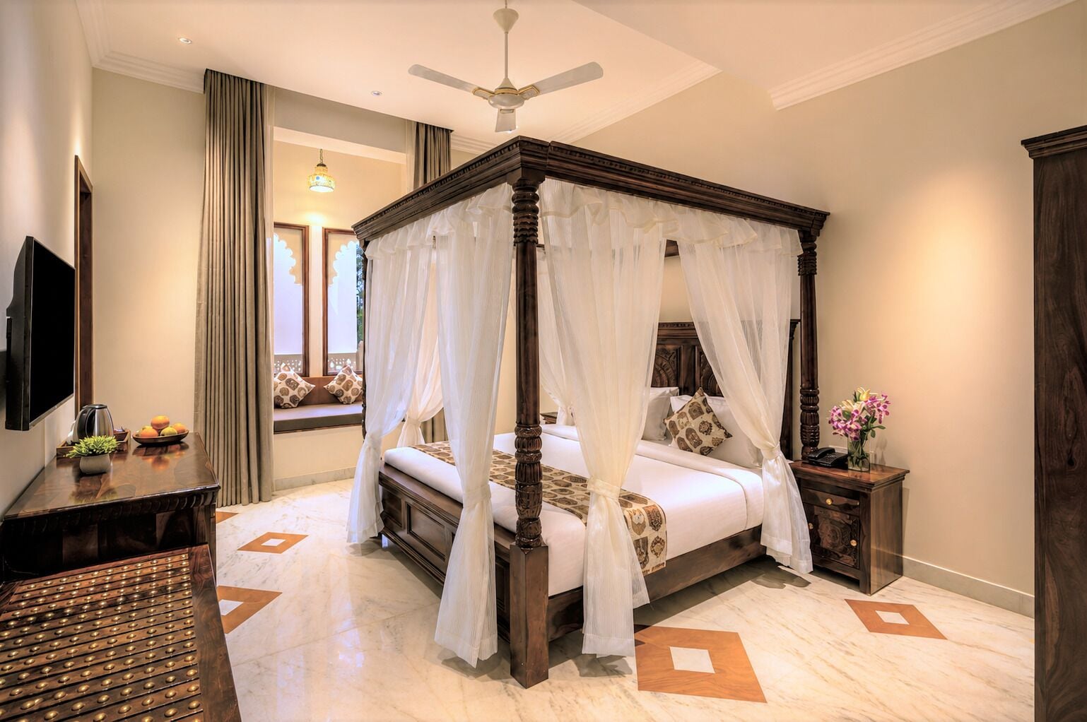A four-poster bed with a canopy, a flat-screen TV, a bedside table with a vase of flowers, and a decorative rug.
