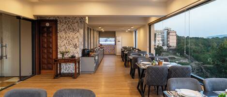 A modern dining area with a long counter, multiple tables, and a view of a cityscape through large windows.