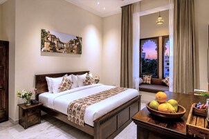 Un cuarto de hotel con una cama grande, una mesita de noche, una mesa de madera con fruta, vistas al atardecer a través de la ventana y un cuadro en la pared.