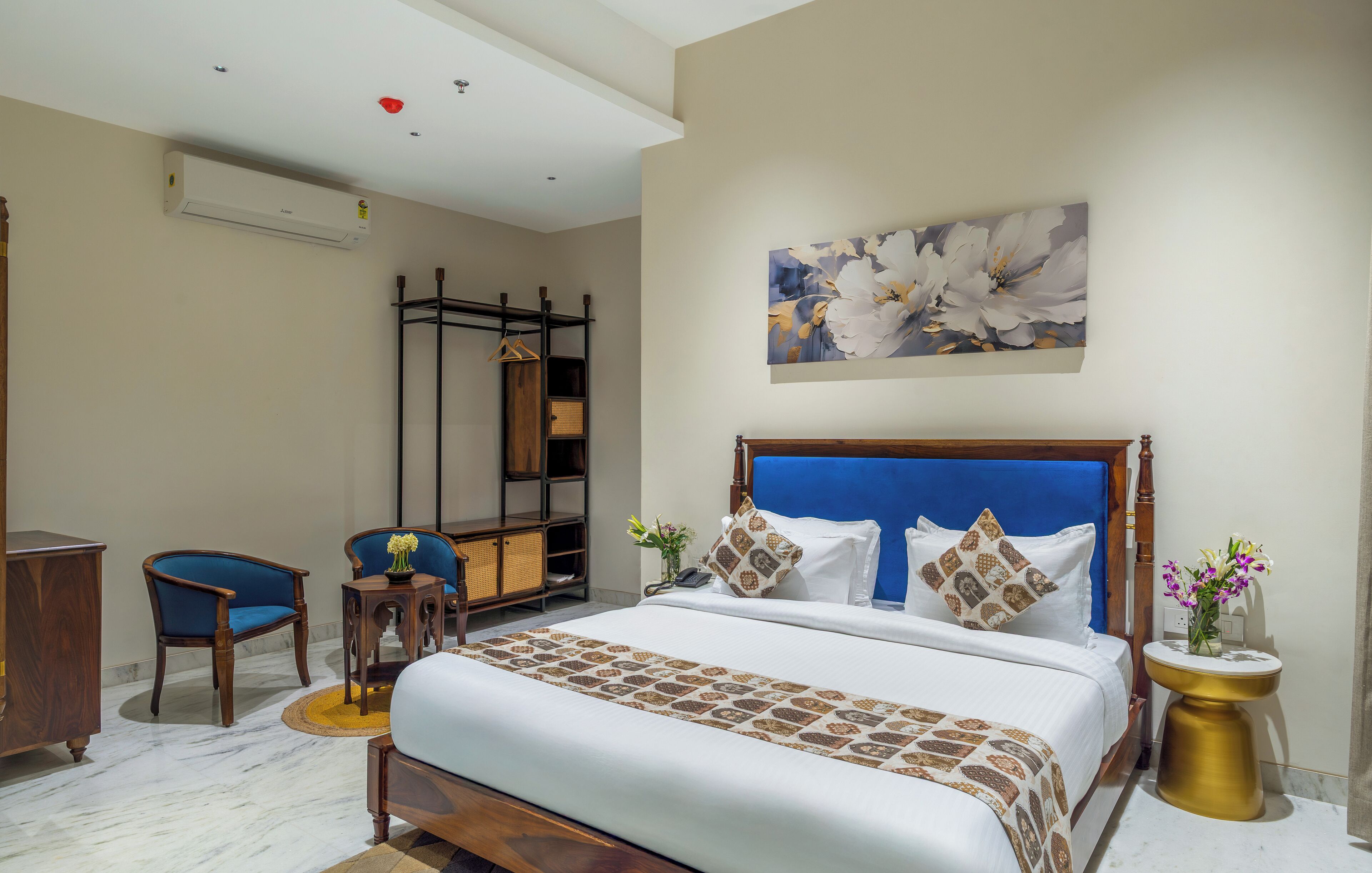 A hotel room with a large bed, a blue upholstered chair, a wooden side table, and a wall-mounted air conditioning unit.