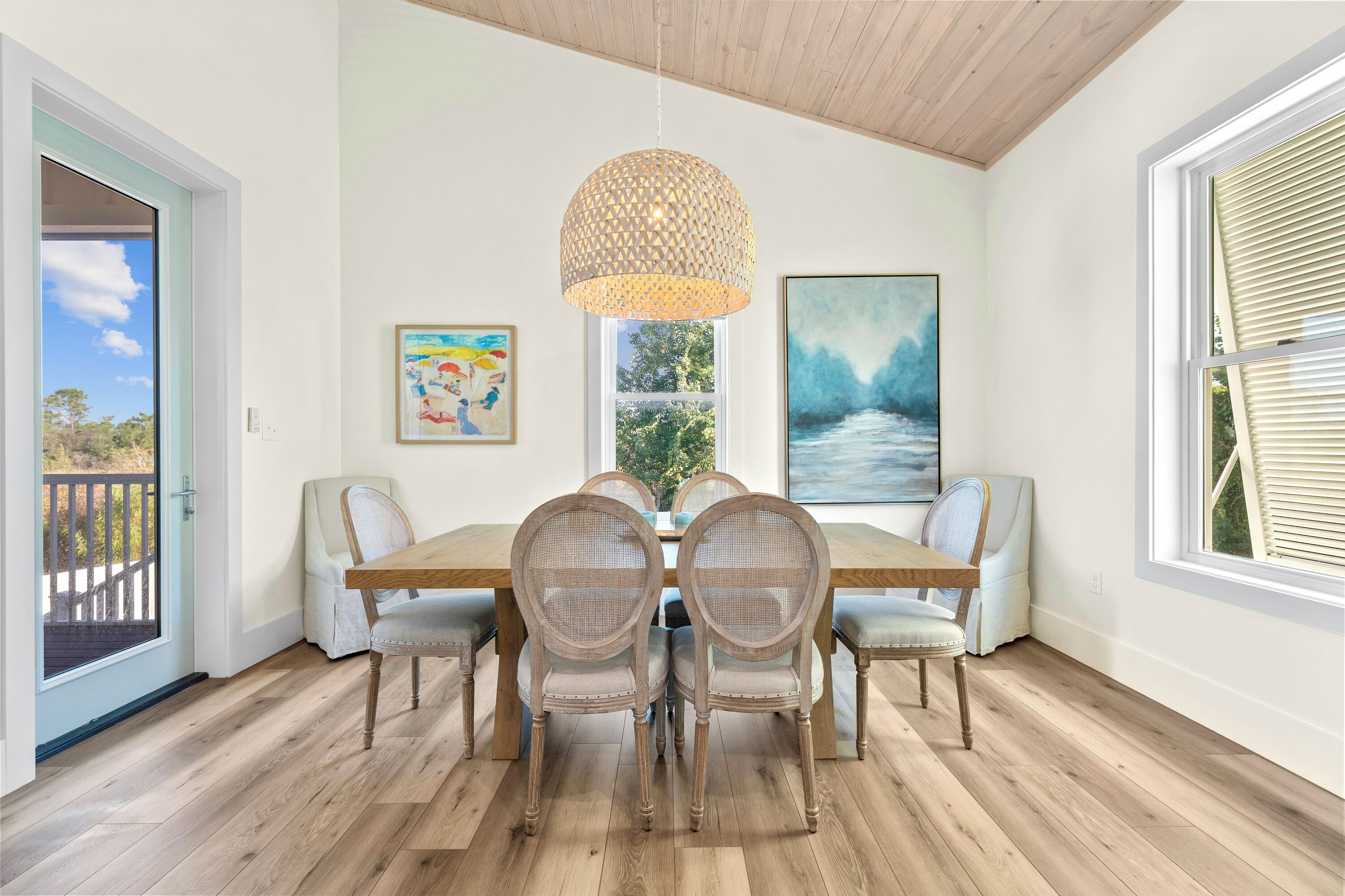 A dining room with a wooden table, wicker chairs, a hanging light fixture, and artwork on the walls.