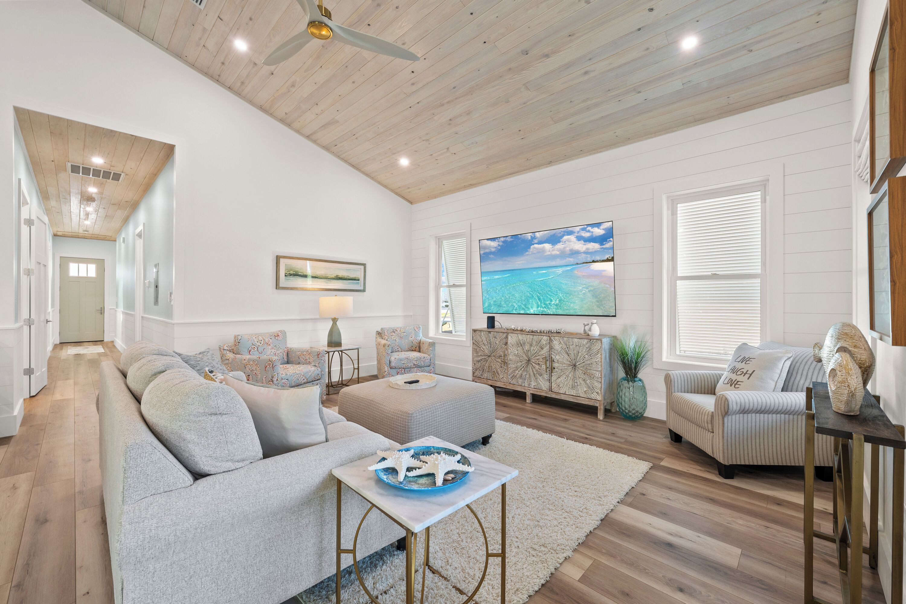A spacious living room with a high ceiling, wooden beams, a large TV, and a view of the ocean through the window.
