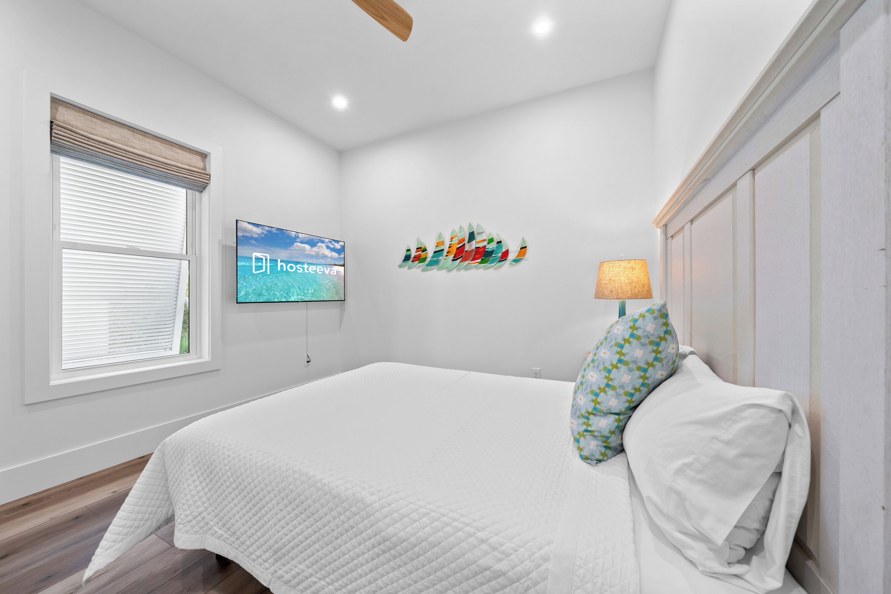 A bedroom with a bed, a TV mounted on the wall, a window with blinds, and a decorative wall piece.