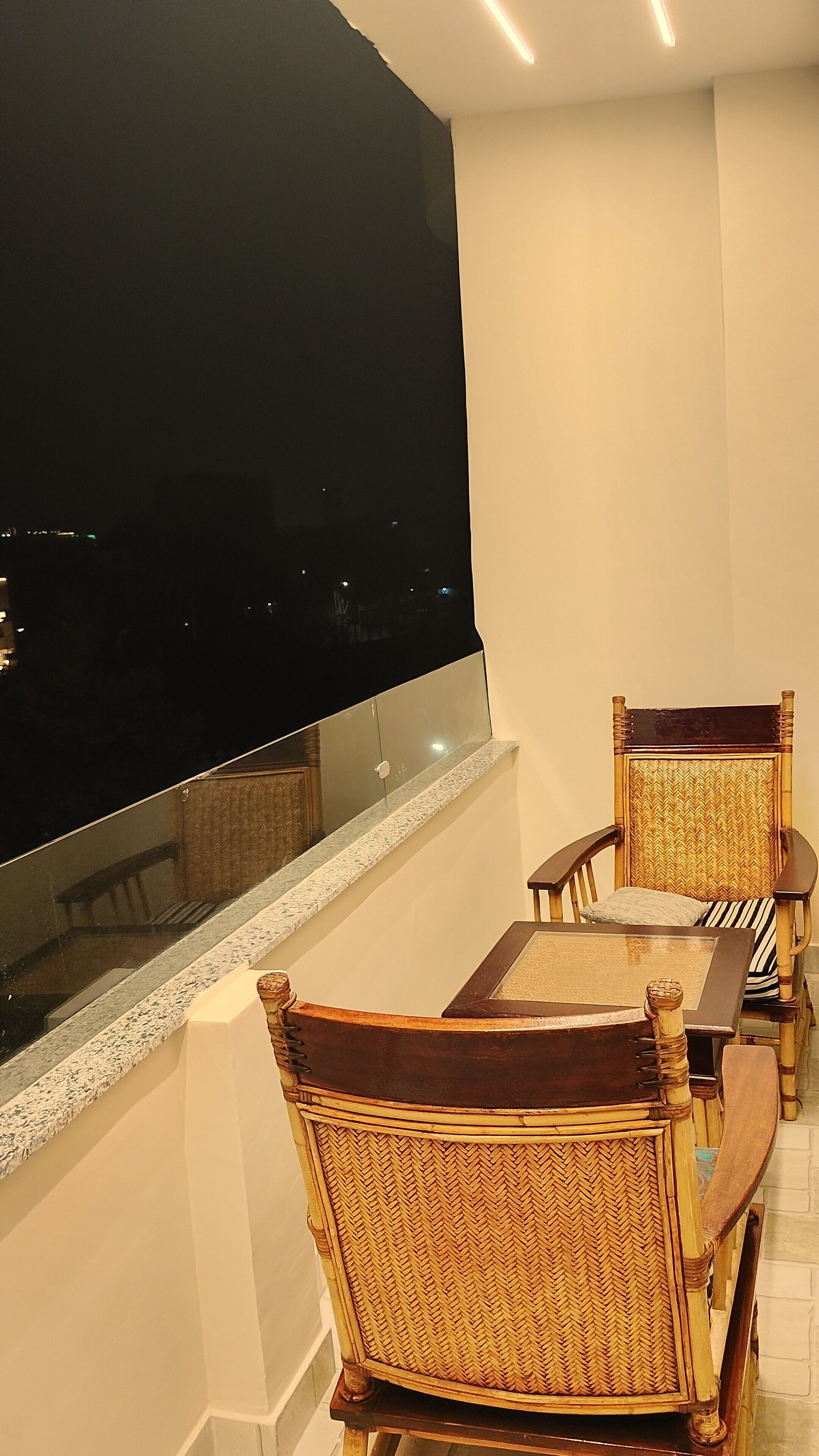A balcony with wicker furniture and a glass railing overlooking a cityscape at night.