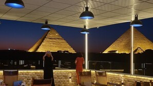 An outdoor seating area with wicker chairs and tables, a brick wall, and a view of pyramids at dusk. - Nefri Pyramids Hotel (Giza)