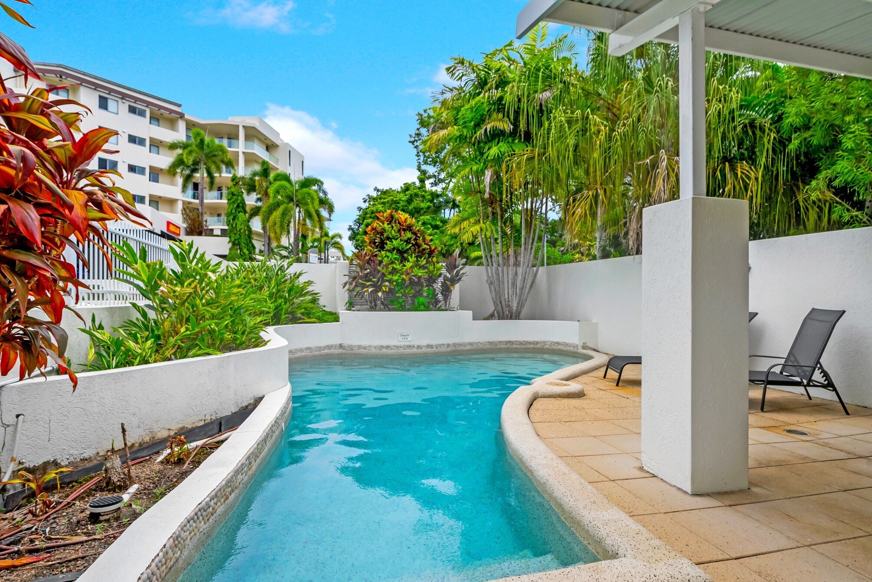 Una piscina con plataforma blanca, rodeada de plantas tropicales y un edificio blanco al fondo.