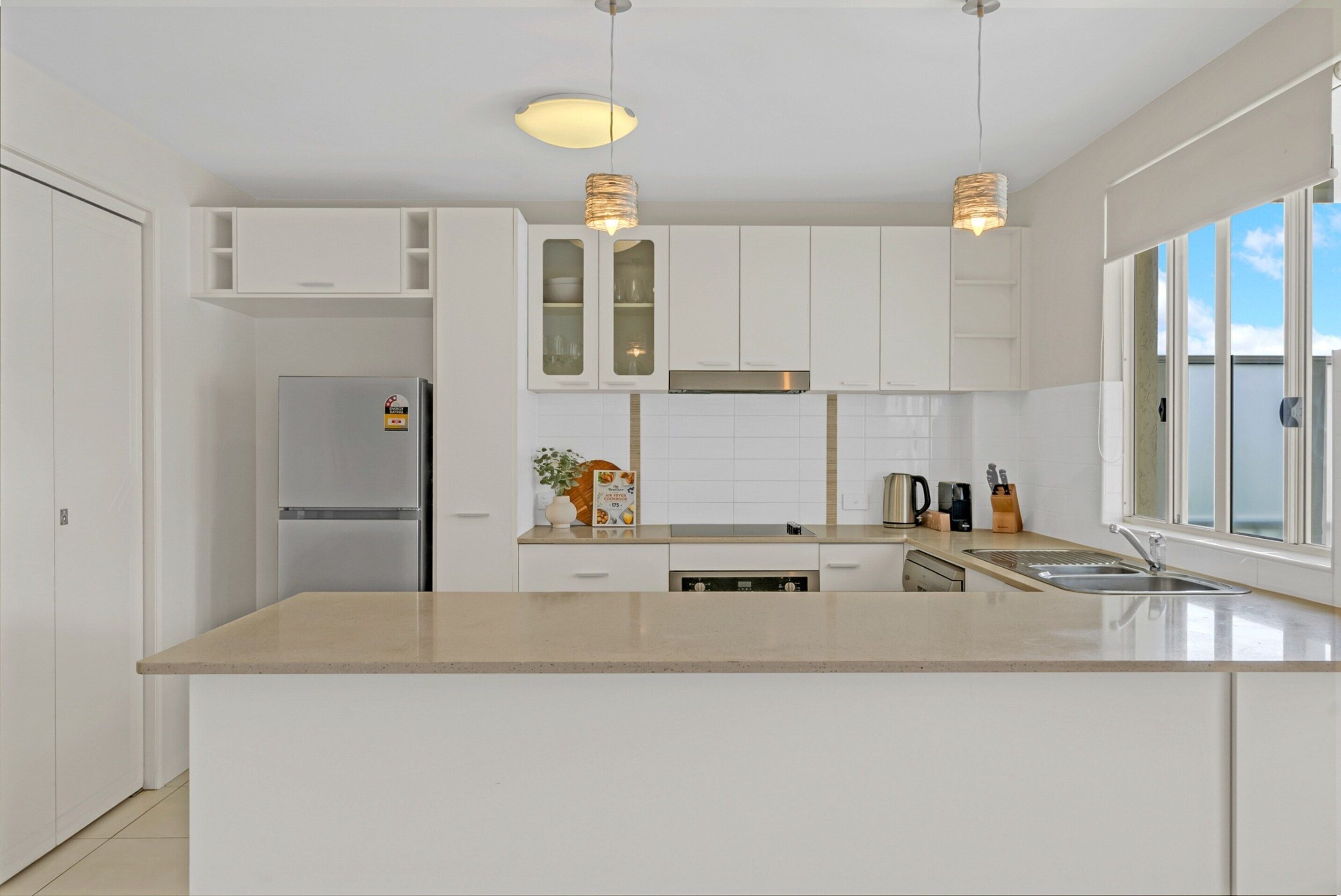 A modern kitchen with white cabinets, a stainless steel refrigerator, a built-in oven, a large island with a sink, and pendant lighting.