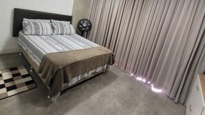 Un dormitorio con cama, ventilador de pie, alfombra a cuadros y un ventanal con cortinas.