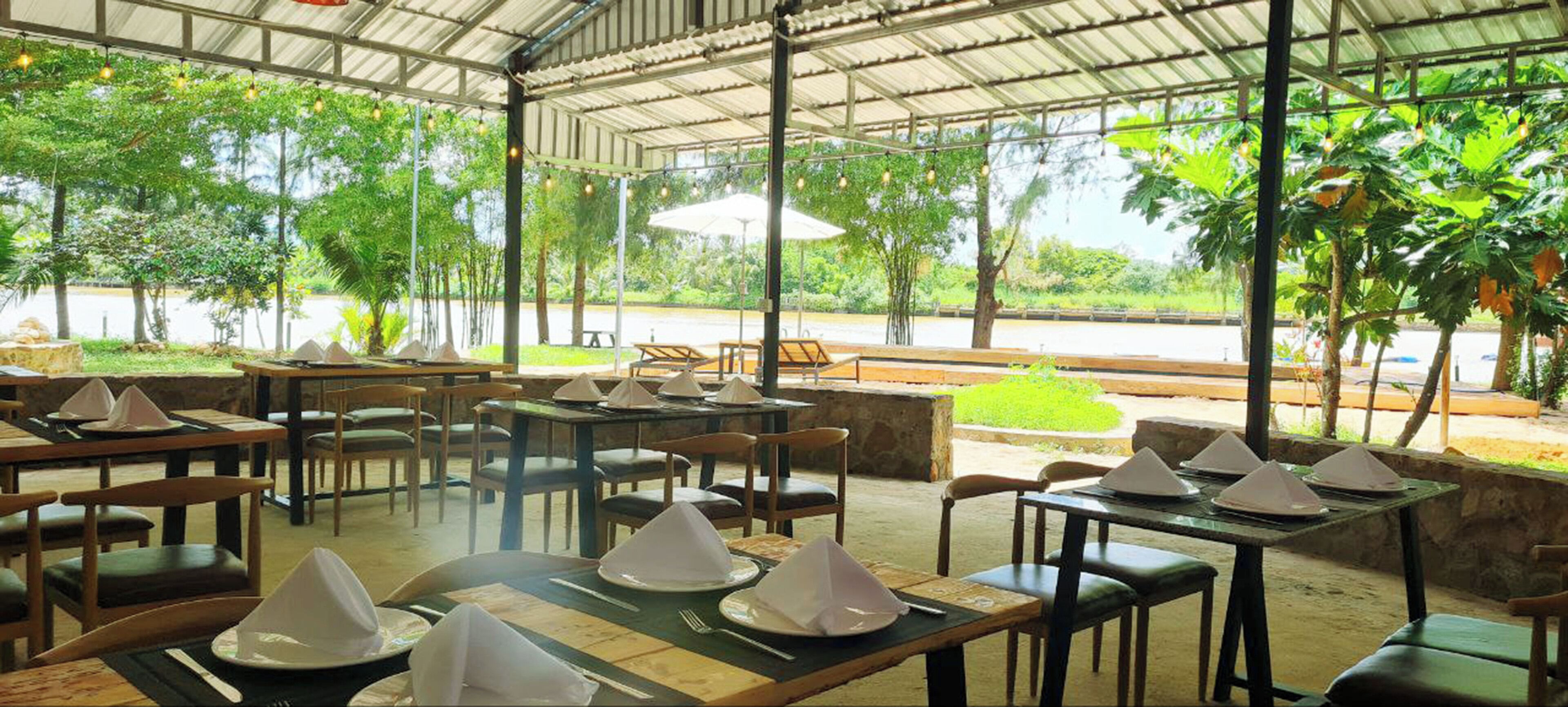 An outdoor restaurant with tables set for dining, surrounded by greenery and a covered area with a view of the road.
