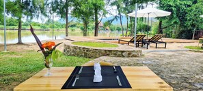 A dining table with a vase of flowers, a set of cutlery, and a napkin. There are two lounge chairs and an umbrella in the background. - Sun&Roots Lodge Restaurant and Bar (Kampot)