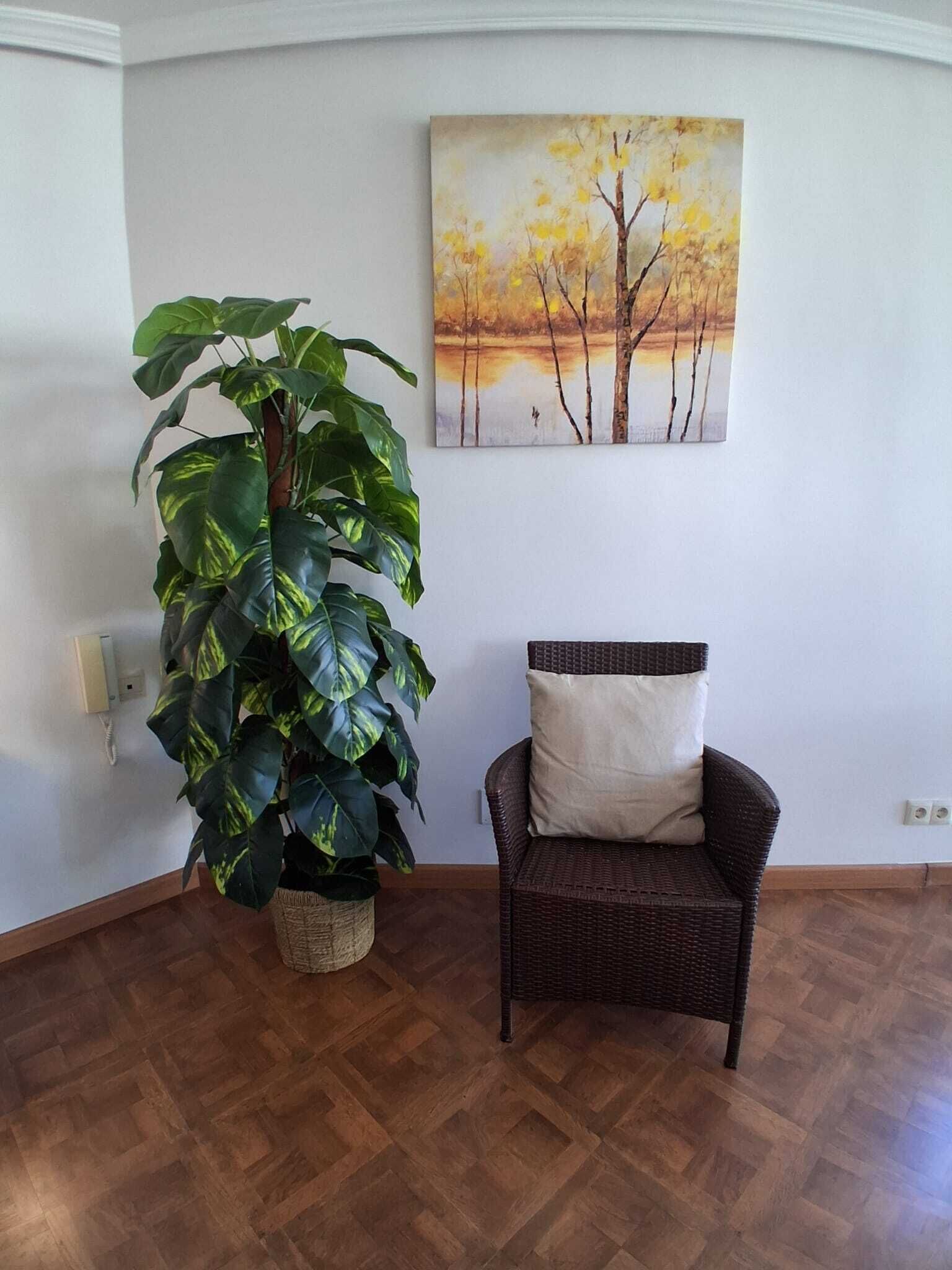 A room with a large potted plant, a wicker chair with a cushion, and a painting of trees on the wall.