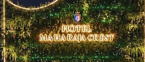 Exterior do Hotel Maharaaja Crest, com uma placa chamativa, um muro decorativo e uma fonte.