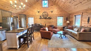 Acogedor interior de cabaña con paredes de madera, un comedor, una sala de estar con sofás y una cocina con refrigerador y mesa de comedor.