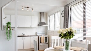 A modern kitchen with white cabinets, a stove, and a sink. There is a dining table with a white tablecloth and woven placemats. A vase with white flowers is on the table. - Luxury Two-Bedroom Apartment (Upper Hutt)