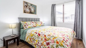 A bedroom with a floral bedspread, a grey headboard, a bedside table with a lamp, and a window with grey curtains. - Luxury Two-Bedroom Apartment (Upper Hutt)