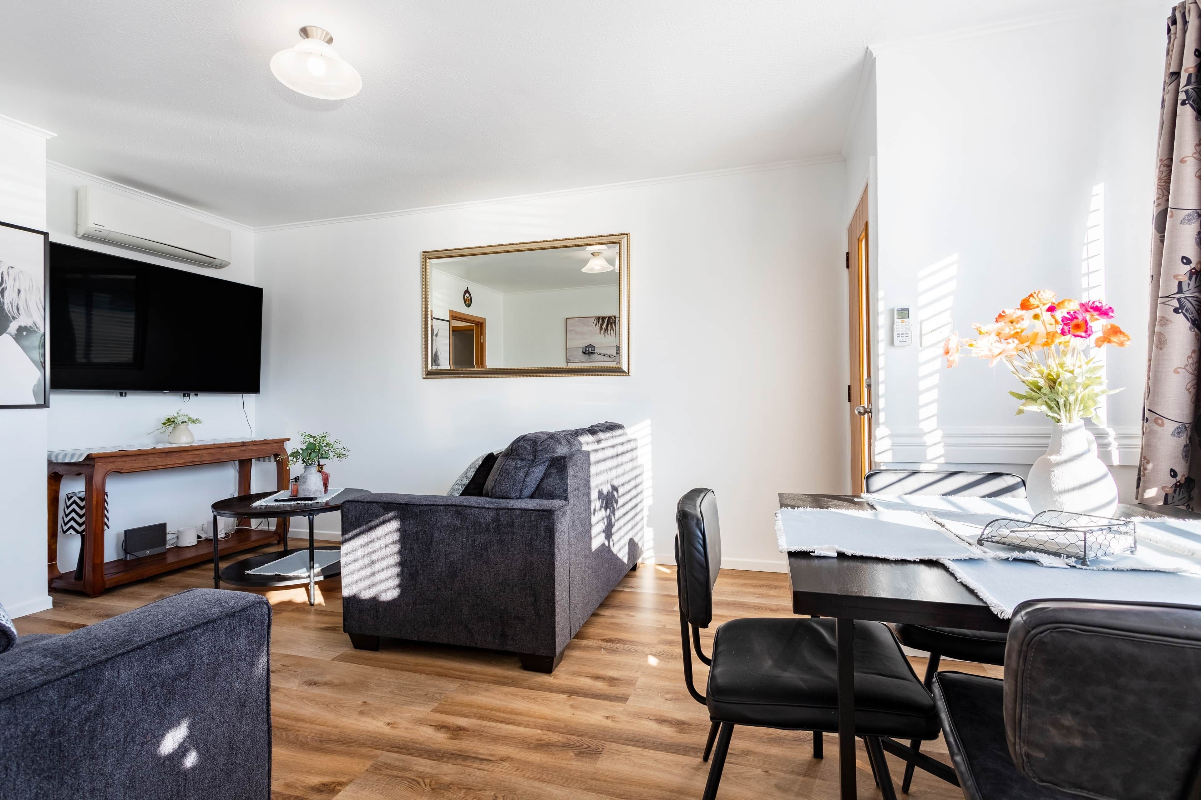 A modern living room with a sofa, a dining table with chairs, a flat-screen TV mounted on the wall, and a mirror above the sofa.