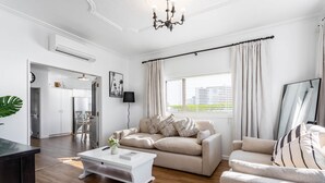 A modern living room with a beige sofa, a white coffee table, and a striped armchair. There is a large window with curtains, a flat-screen TV, and a chandelier. - Deluxe Three Bedroom Villa (Upper Hutt)