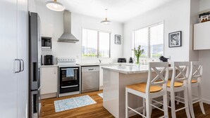A modern kitchen with white cabinetry, stainless steel appliances, and a wooden dining area with white chairs. - Deluxe Three Bedroom Villa (Upper Hutt)