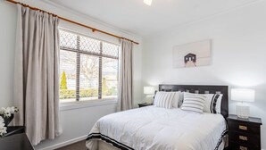 A bedroom with a large bed, a nightstand with a lamp, a window with curtains, and a framed picture on the wall. - Deluxe Three Bedroom Villa (Upper Hutt)