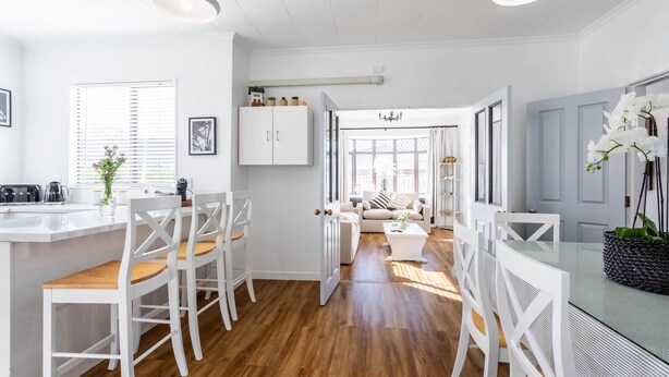 A modern kitchen with white cabinetry, wooden flooring, and a central island with bar stools. - Deluxe Three Bedroom Villa (Upper Hutt)