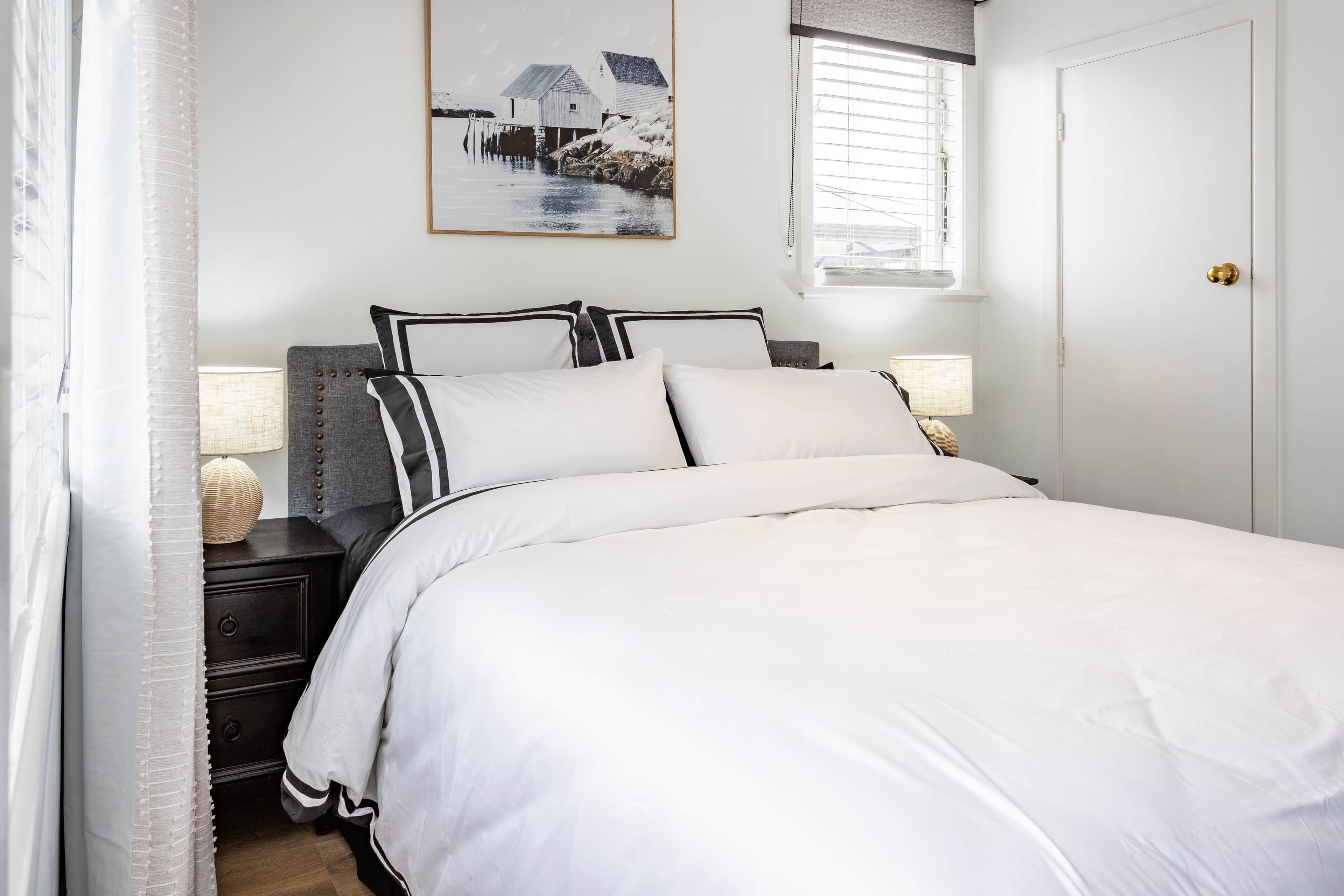 A neatly made bed with white bedding, a bedside table with a lamp, and a framed picture on the wall.