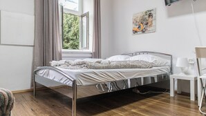 Een slaapkamer met een metalen bedframe, een witte nachtkastje met een lamp, een raam met gordijnen en een aan de muur bevestigde televisie.