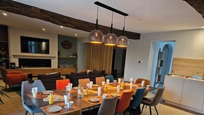 A dining area with a large table set for six, surrounded by modern chairs. There is a television mounted on the wall, a wooden beam ceiling, and a kitchen area visible in the background. - A la Gloriette Futuroscope (Neuville-de-Poitou)
