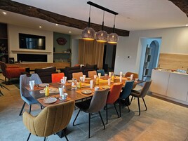 Uma área de jantar com uma mesa grande posta para seis pessoas, rodeada por cadeiras modernas. Há uma televisão montada na parede, teto com vigas de madeira e uma cozinha visível ao fundo.
