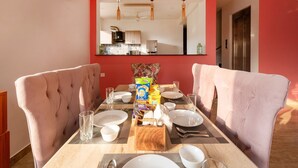 Un comedor con una mesa preparada para cuatro, dos sillas acolchadas, una cocina al fondo y un equipo de aire acondicionado en la pared.